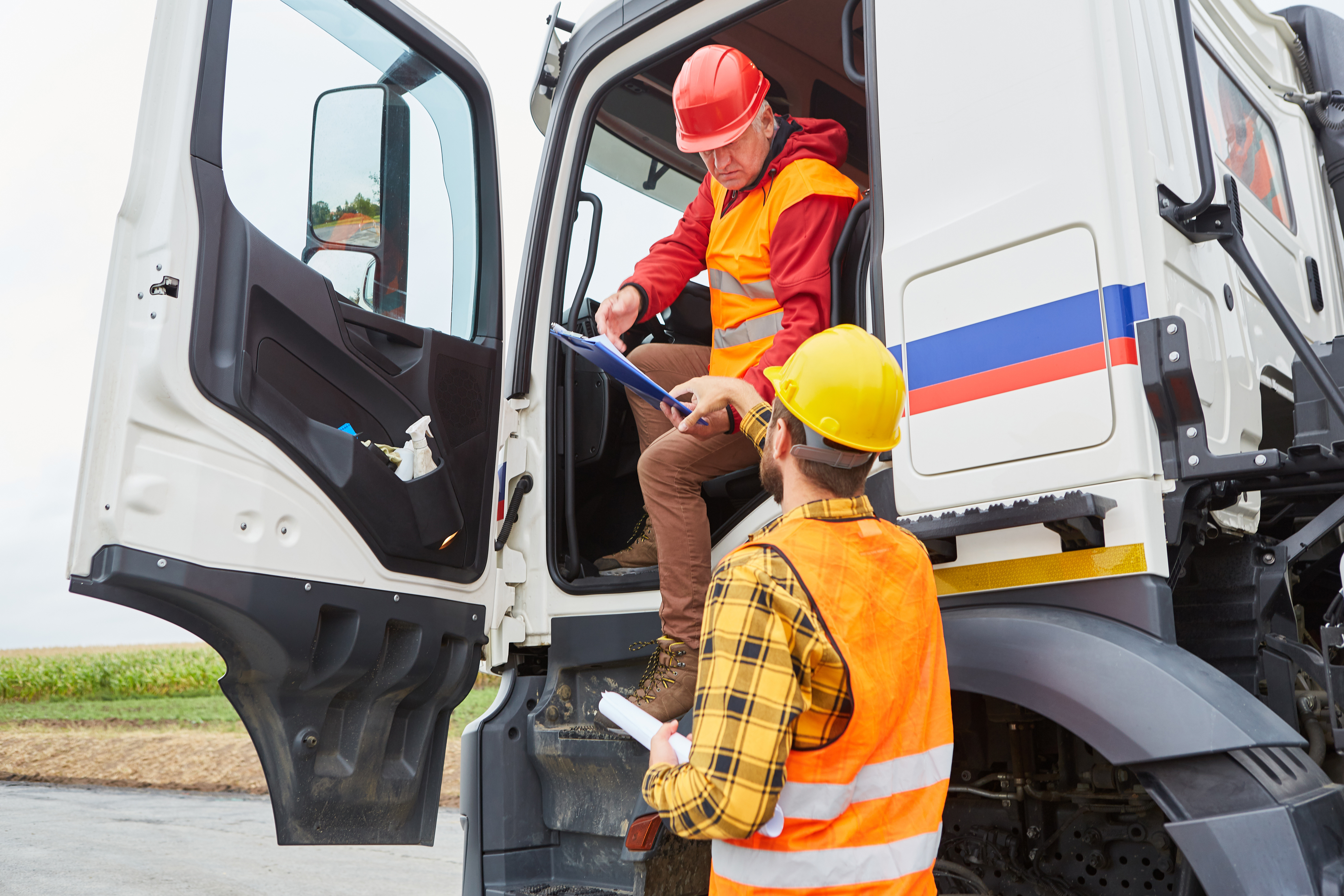 Deux chauffeurs de camion en gilets de sécurité et casques (un assis dans la cabine du camion, l'autre debout à l'extérieur) discutent une liste de contrôle.