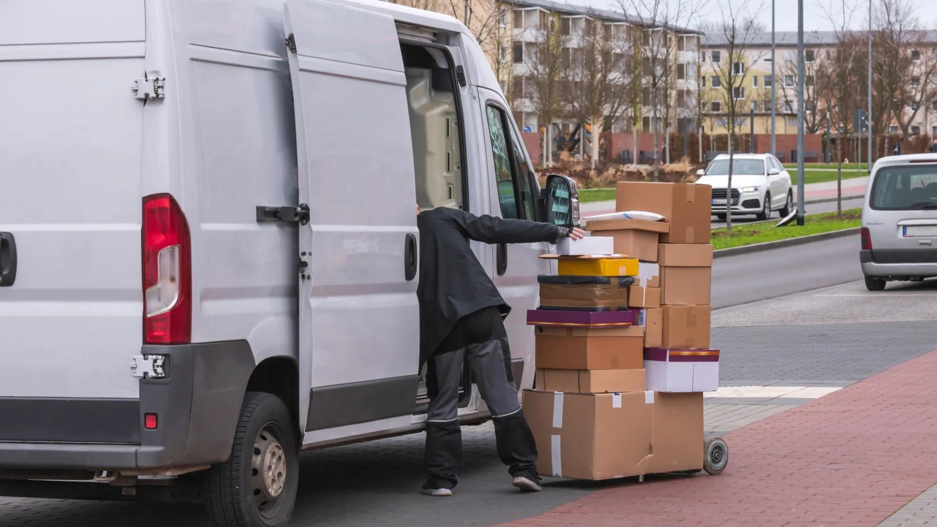 A delivery person unloads stacked cardboard boxes from a white van parked on a brick street in a residential area