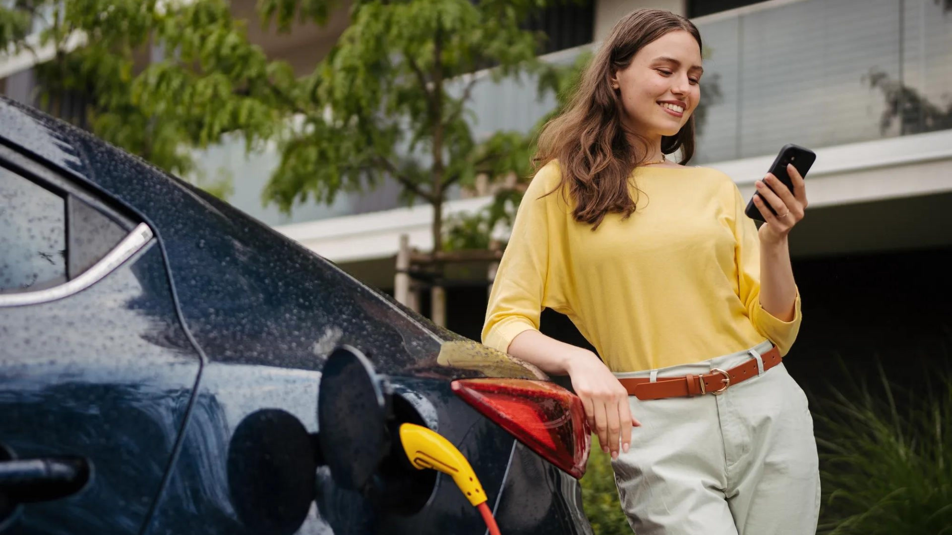 Woman in yellow shirt holding phone while charging electric car with yellow connector cable