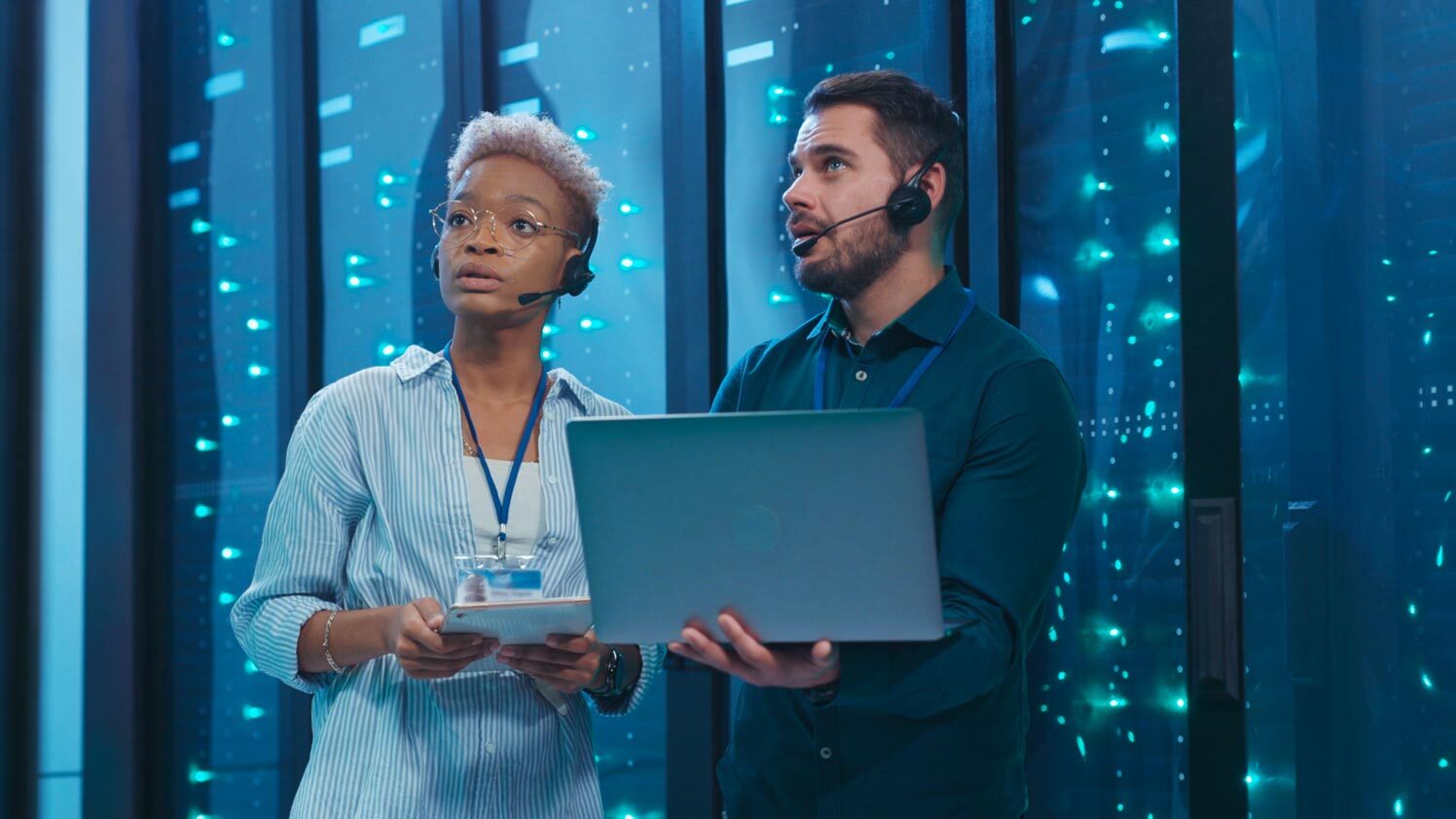 Two IT security engineers reviewing data on a laptop inside a server room.