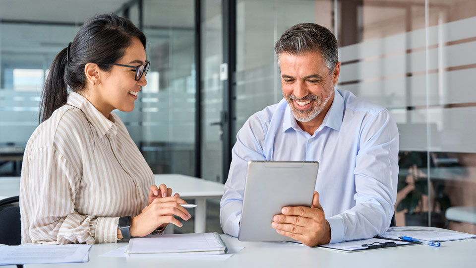 Two people sat at a desk looking at a tablet and smiling