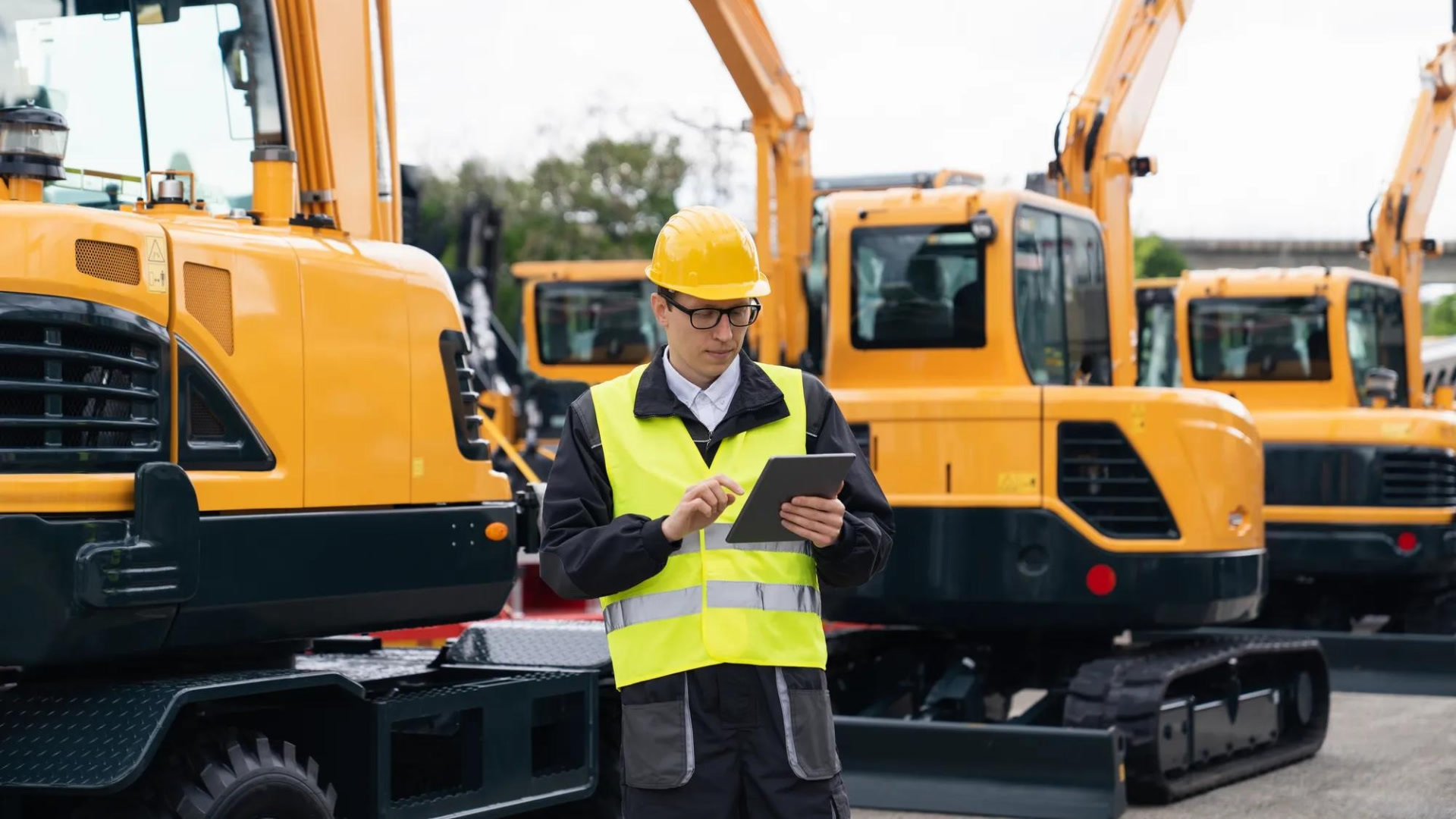 Un uomo con elmetto giallo e gilet catarifrangente utilizza un tablet per la gestione della flotta davanti a degli escavatori gialli