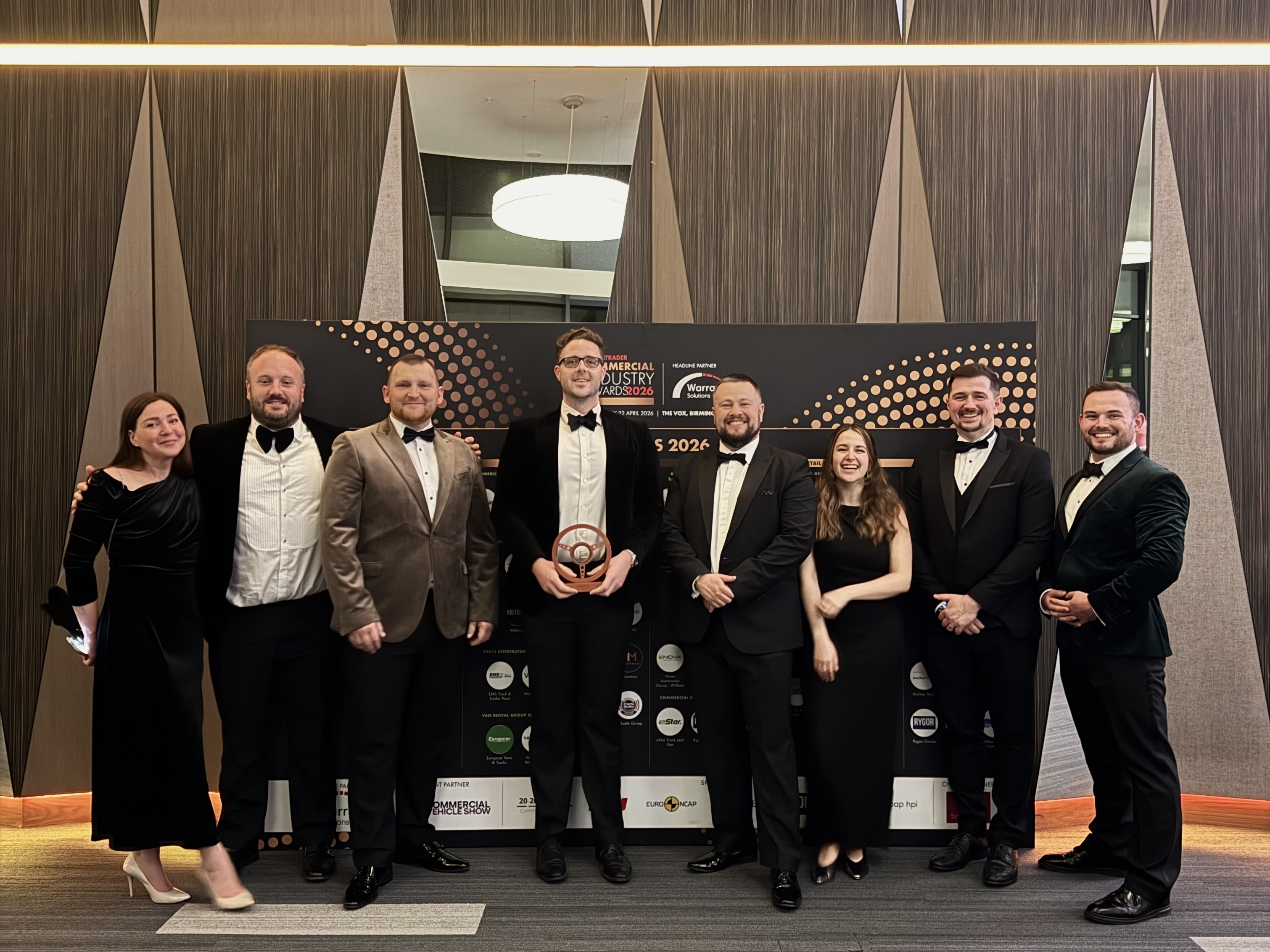 A group of eight people dressed in formal evening wear posing together in front of a Motor Trader Commercial Industry Awards 2026 backdrop, with one person in the center holding the wooden award.