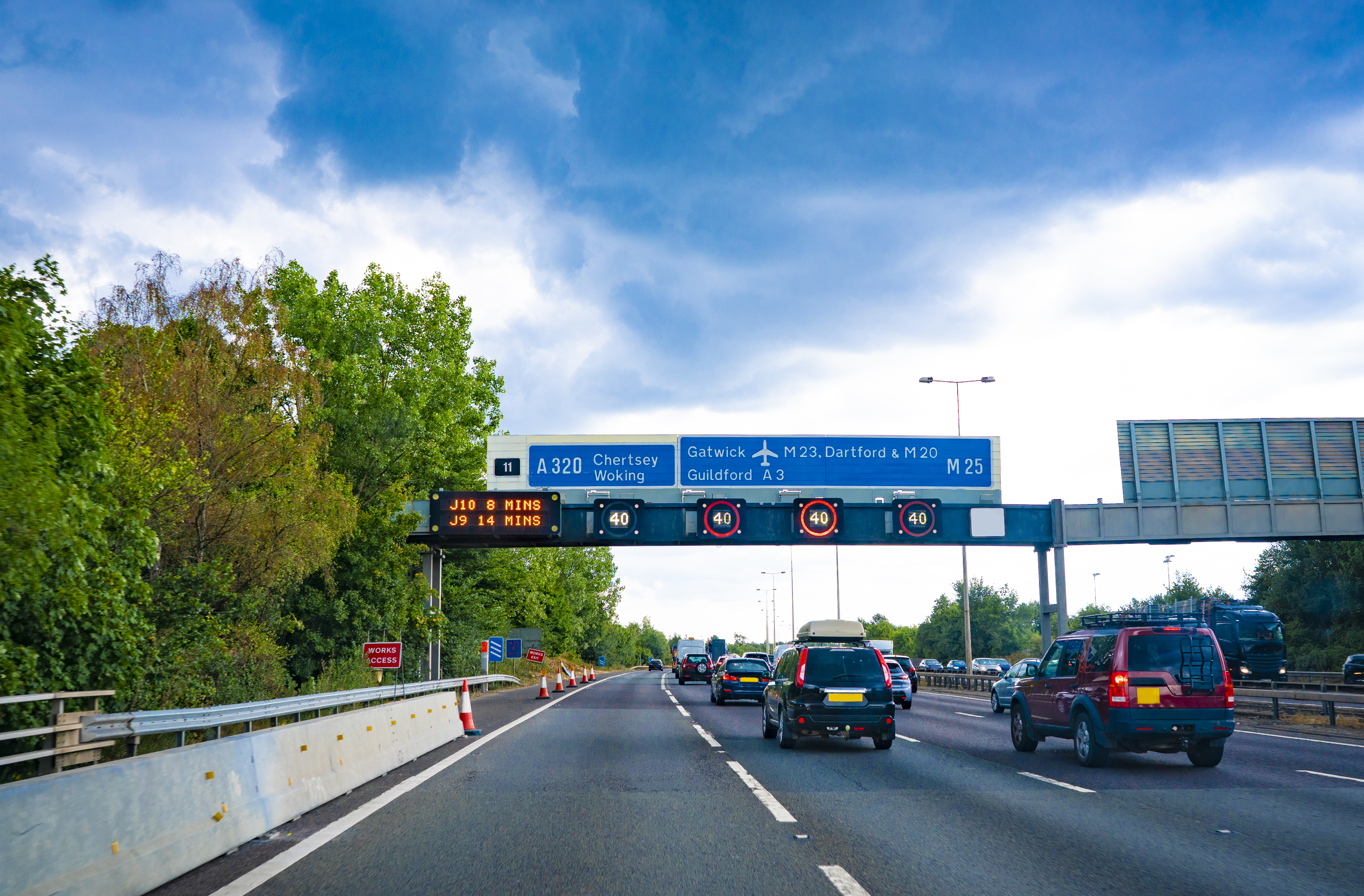 Traffic on a highway under an overhead gantry displaying a 40 speed limit, travel times to Junctions 9 and 10, and signs for M25, Gatwick, and Chertsey