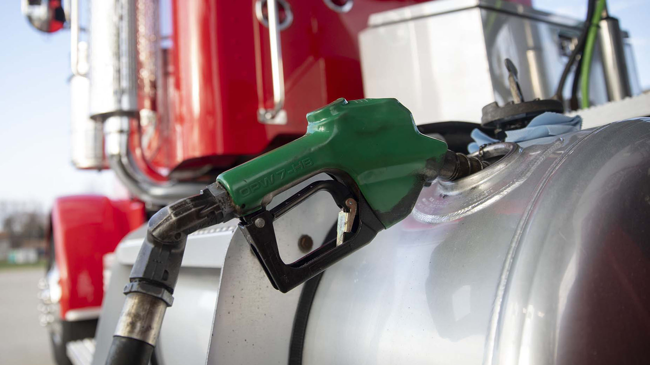 Uma bomba de combustível verde está conectada ao tanque de um caminhão de carga vermelho, ilustrando o momento do abastecimento. A cena destaca a importância do controle preciso de consumo e eficiência de combustível na operação de frotas pesadas.