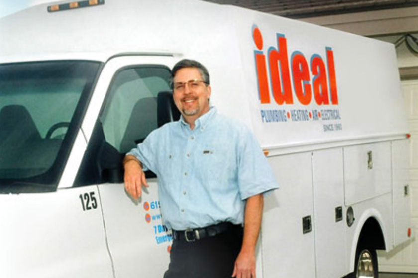 A smiling man in a blue work shirt stands up against a white Ideal work van