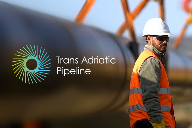 Employee wearing orange safety vest and white hard helmet standing next to a large pipe.