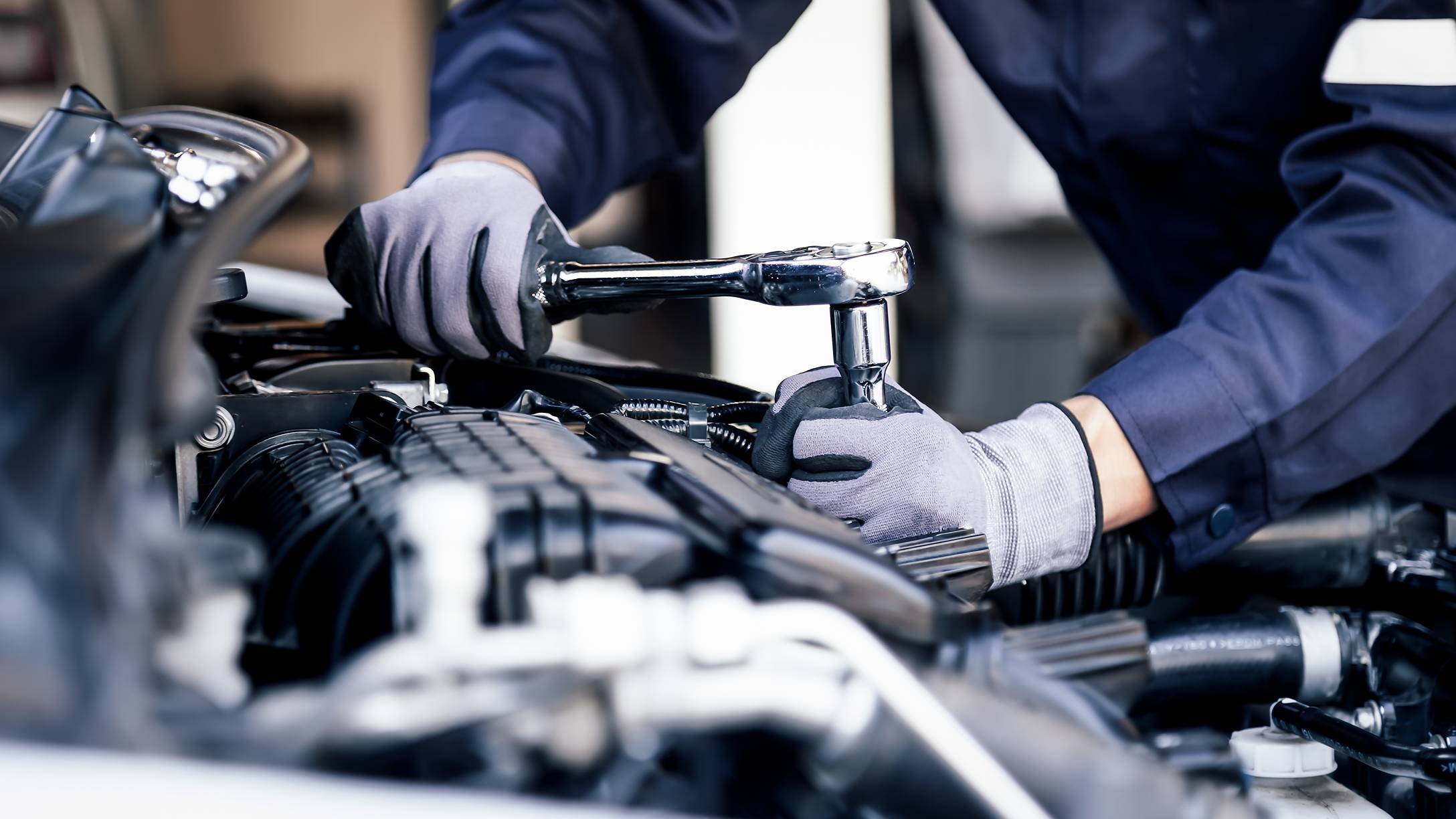 Nahaufnahme eines Mechanikers in Bluse und Handschuhen, der mit einer Ratsche an einem Fahrzeugmotor arbeitet.